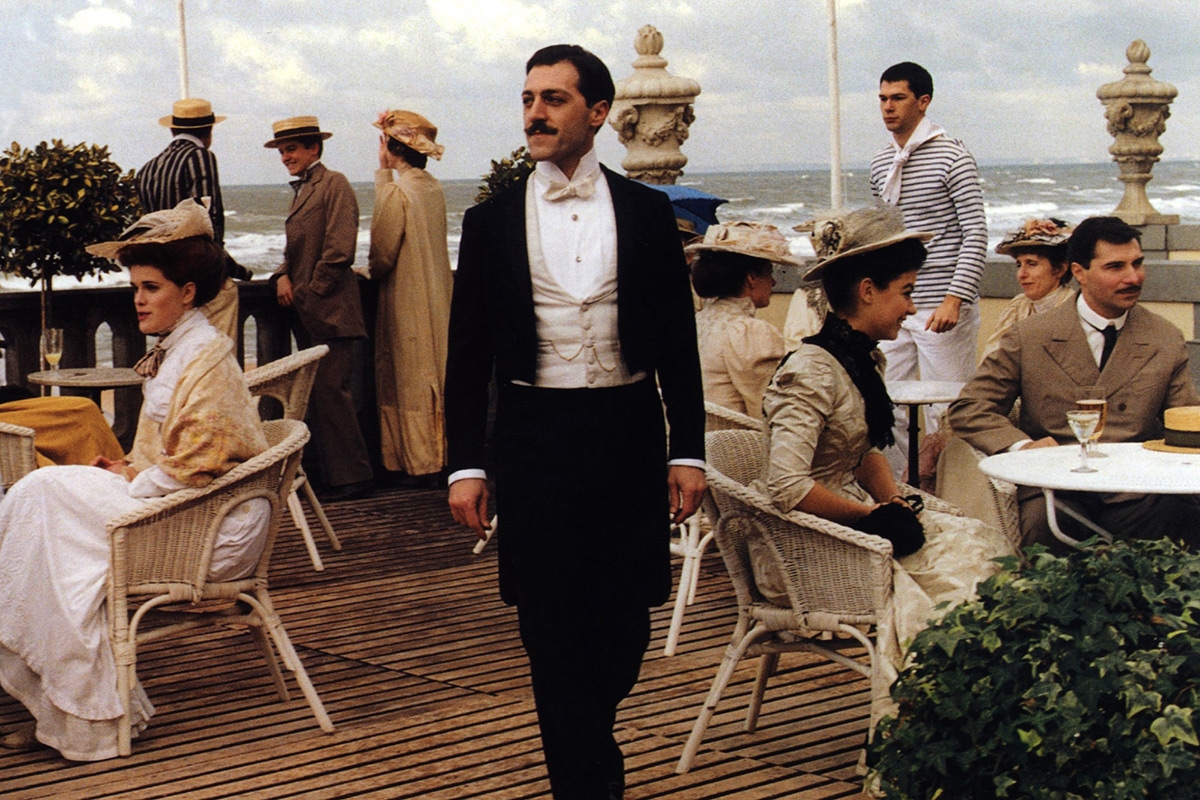 Filmstill aus »Die wiedergefundene Zeit«, Marcel Proust läuft zwischen Tischen einer Außengastronomie im Hintergrund sieht man das Meer 