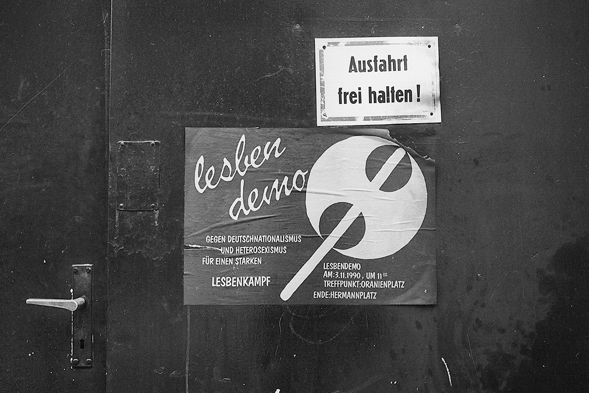 Schwarze Wand mit einem Plakat, auf dem eine Doppelaxt zu sehen ist, daneben der Schriftzug »lesbendemo«, darunter »Gegen Deutschnationalismus und Heterosexismus, für einen starken Lesbenkampf. Lesbendemo, Am 3.11.1990, um 11.00, Treffpunkt: Oranienplatz, Ende: Hermannplatz«