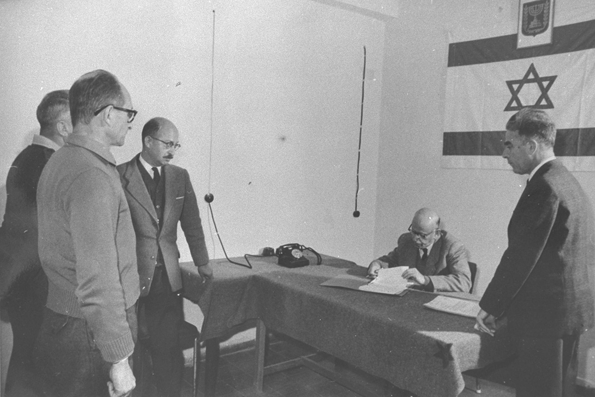 Der Untersuchungsrichter (sitzend) genehmigt den Antrag auf Verlängerung der Vernehmung von Eichmann (links vorn) im Beisein von Elrom (r.), Jerusalem, 9. März 1961