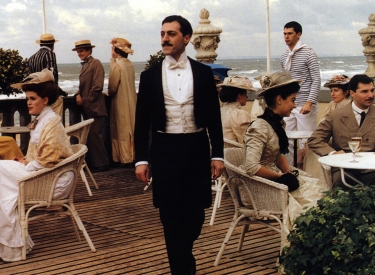 Filmstill aus »Die wiedergefundene Zeit«, Marcel Proust läuft zwischen Tischen einer Außengastronomie im Hintergrund sieht man das Meer 